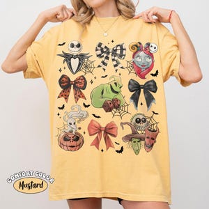 May include: A mustard yellow t-shirt featuring a graphic print of various characters and bows. The design includes Jack Skellington, Sally, Oogie Boogie, Zero, and three trick-or-treaters, interspersed with black and orange bows and spiderwebs.