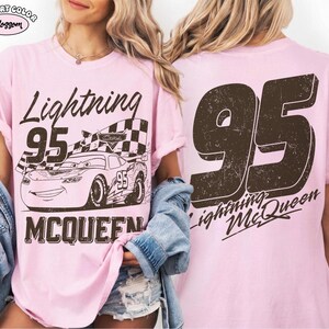 May include: Pink t-shirt featuring a graphic of Lightning McQueen from the movie Cars, with the number 95 and the name McQueen. The back of the shirt displays a large number 95 and the words "Lightning McQueen".