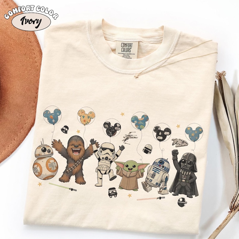 May include: Ivory-colored t-shirt featuring a colorful graphic of Star Wars characters, including BB-8, Chewbacca, a Stormtrooper, Grogu, R2-D2, and Darth Vader. The characters are holding Mickey Mouse-shaped balloons. The shirt has the Comfort Colors brand label.
