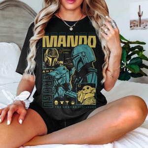 K&ouml;nnte beinhalten: Schwarzes T-Shirt mit einem gelb und t&uuml;rkis farbenen Grafikdesign. Das Design zeigt den Text "MANDO AND GROGU" und Illustrationen eines Mandalorianer-Helms und Grogu, mit dem Satz "THE LEGEND CONTINUES".