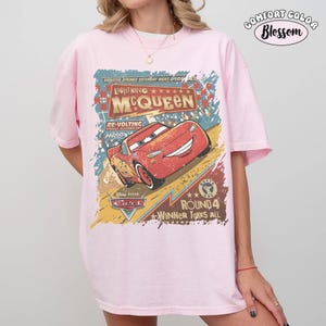 May include: A light pink Comfort Colors t-shirt featuring a distressed graphic of Lightning McQueen from the Disney Pixar Cars movie. The graphic includes text that reads "Lightning McQueen" and "Winner Takes All".