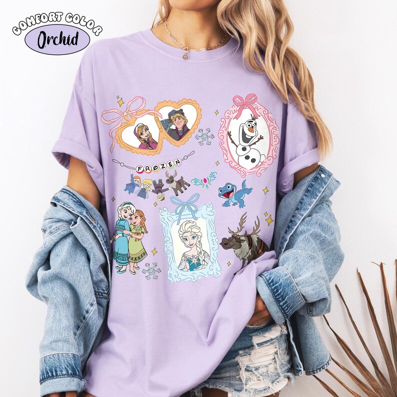 May include: A lilac Comfort Colors t-shirt featuring framed illustrations of characters from the movie Frozen. The shirt includes images of Anna, Elsa, Olaf, Kristoff, Sven, and other characters. The word "FROZEN" is also printed on the shirt.