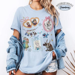 May include: Light blue t-shirt featuring various characters and scenes from the animated movie "Frozen." The design includes images of Anna, Elsa, Olaf, Kristoff, Sven, and other characters, with the word "FROZEN" in the center. The shirt has rolled-up sleeves.