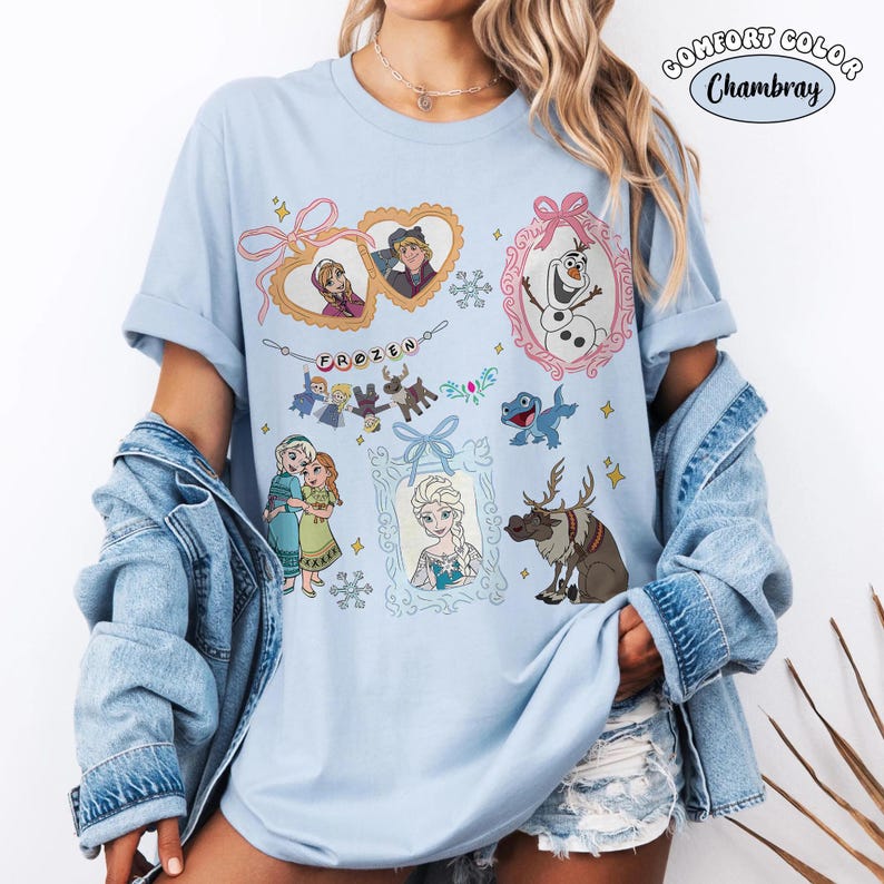 May include: Light blue t-shirt featuring various characters and scenes from the animated movie "Frozen." The design includes images of Anna, Elsa, Olaf, Kristoff, Sven, and other characters, with the word "FROZEN" in the center. The shirt has rolled-up sleeves.