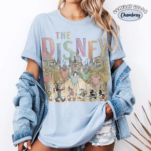 Pu&ograve; includere: T-shirt azzurra con la scritta "THE DISNEY" in colori pastello. La grafica presenta i personaggi Disney che sfilano davanti a un castello, tenendo in mano palloncini di Topolino. La maglietta &egrave; etichettata "Comfort Color Chambray".