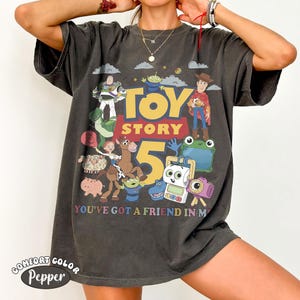 May include: A charcoal gray short-sleeve t-shirt featuring a graphic print of Toy Story characters including Buzz Lightyear, Woody, and Jessie. The design includes the text TOY STORY 5 and YOU'VE GOT A FRIEND IN ME, surrounded by various colorful toy figures.