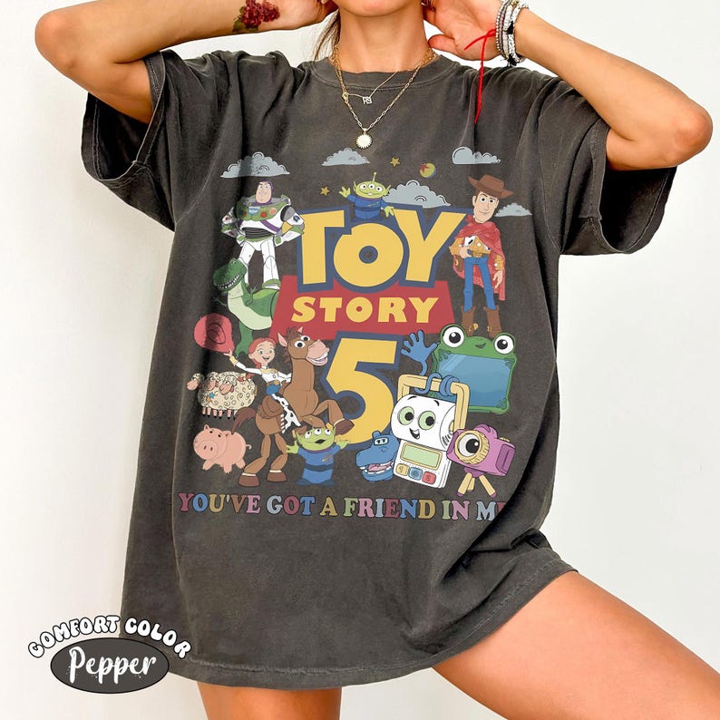May include: A charcoal gray short-sleeve t-shirt featuring a graphic print of Toy Story characters including Buzz Lightyear, Woody, and Jessie. The design includes the text TOY STORY 5 and YOU'VE GOT A FRIEND IN ME, surrounded by various colorful toy figures.
