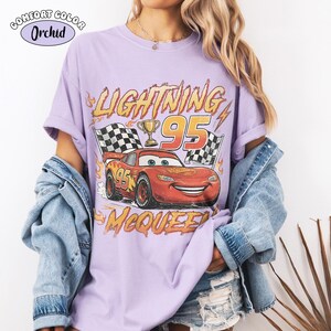 Puede incluir: Una camiseta de color morado claro con un gr&aacute;fico de un coche de carreras rojo con el n&uacute;mero 95, un trofeo y banderas a cuadros. El texto dice LIGHTNING y MCQUEEN. Una etiqueta en la esquina superior izquierda muestra COMFORT COLOR y ORCHID.