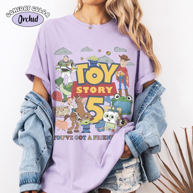 May include: A light purple t-shirt featuring a graphic print of Toy Story characters including Woody, Buzz Lightyear, and Rex. The design includes the text TOY STORY 5 and YOU'VE GOT A FRIEND. The shirt is labeled as Comfort Color Orchid.