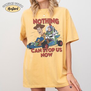 May include: Mustard-colored t-shirt with a graphic featuring Woody and Buzz Lightyear in a vehicle, with the text "NOTHING CAN STOP US NOW" in red. The shirt has short sleeves and a relaxed fit.