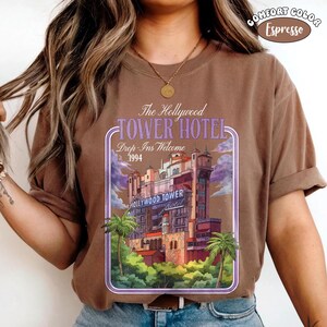 Op de afbeelding: Bruin T-shirt met een afbeelding van het Hollywood Tower Hotel, met de tekst "The Hollywood Tower Hotel, Drop-Ins Welcome, 1994." Het ontwerp is omlijst door een paarse rand met palmbomen.