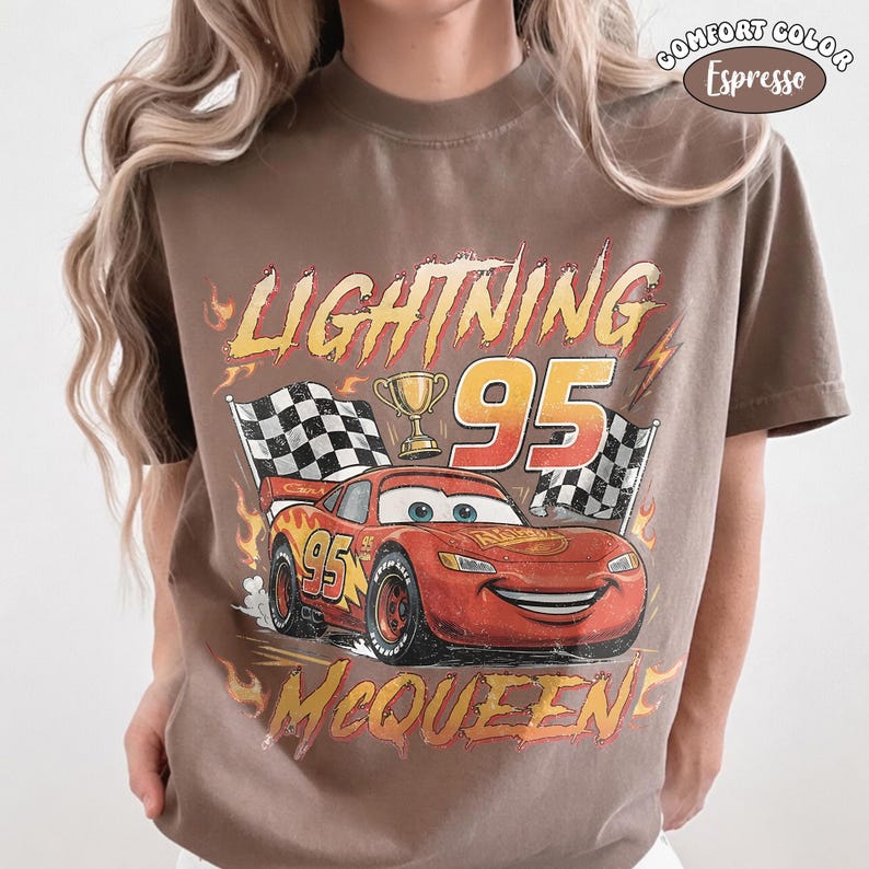 Puede incluir: Una camiseta color marr&oacute;n espresso con un gr&aacute;fico del coche de carreras rojo Lightning McQueen, el n&uacute;mero 95, un trofeo dorado y banderas a cuadros. El texto dice LIGHTNING MCQUEEN y 95, rodeado de llamas naranjas y una etiqueta Comfort Color.