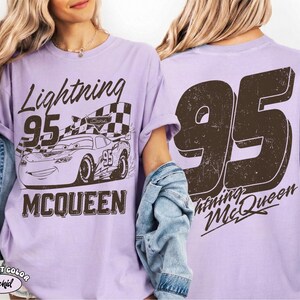 May include: A light purple t-shirt featuring a graphic of Lightning McQueen race car with the number 95 and the text "Lightning McQueen". The back of the shirt displays a large number 95 and "Lightning McQueen".