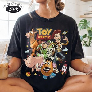 May include: Black Comfort Colors t-shirt featuring a colorful graphic of Toy Story characters, including Woody, Buzz Lightyear, and Mr. Potato Head. The Toy Story logo is displayed above the characters. The shirt is a crew neck style.
