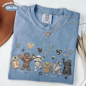 May include: A blue Comfort Colors t-shirt featuring Star Wars characters, including BB-8, Chewbacca, a Stormtrooper, Grogu, R2-D2, and Darth Vader. The shirt has a graphic of the characters with Mickey Mouse-shaped balloons. The shirt also has the text "Blue Jean".