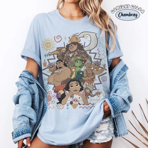 Puede incluir: Una camiseta azul claro con un estampado gr&aacute;fico de personajes de la pel&iacute;cula Moana, incluyendo a Maui, Moana, Heihei, Pua y la abuela Tala. El dise&ntilde;o incluye patrones tribales y un anzuelo. El texto Comfort Color Chambray es visible.