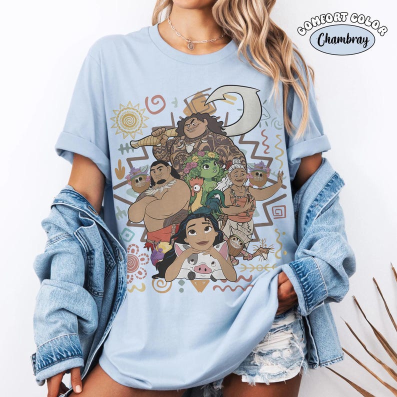 Puede incluir: Una camiseta azul claro con un estampado gr&aacute;fico de personajes de la pel&iacute;cula Moana, incluyendo a Maui, Moana, Heihei, Pua y la abuela Tala. El dise&ntilde;o incluye patrones tribales y un anzuelo. El texto Comfort Color Chambray es visible.