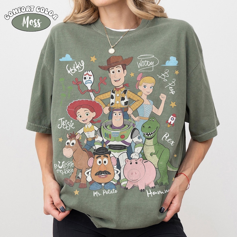 Może przedstawiać: Mchowo-zielony t-shirt z kr&oacute;tkim rękawem z nadrukiem graficznym postaci z Toy Story, w tym Woody'ego, Buzza Astrala, Jessie, Rexa, Pana Bulwy, Celka, Sztućka i Pastereczki, z ich imionami wypisanymi białym pismem odręcznym.