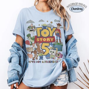 May include: A light blue short-sleeve t-shirt featuring a graphic print of Toy Story characters including Woody, Buzz Lightyear, and Rex. The text reads Toy Story 5 and You've Got A Friend In Me. A small circular logo in the corner says Comfort Color Chambray.