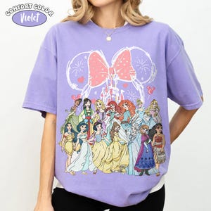 Op de afbeelding: Een violet Comfort Colors T-shirt met een afbeelding van Disney-prinsessen. Het ontwerp bevat een roze strik, vuurwerk en een kasteelomtrek. Het shirt heeft korte mouwen en een ronde hals.