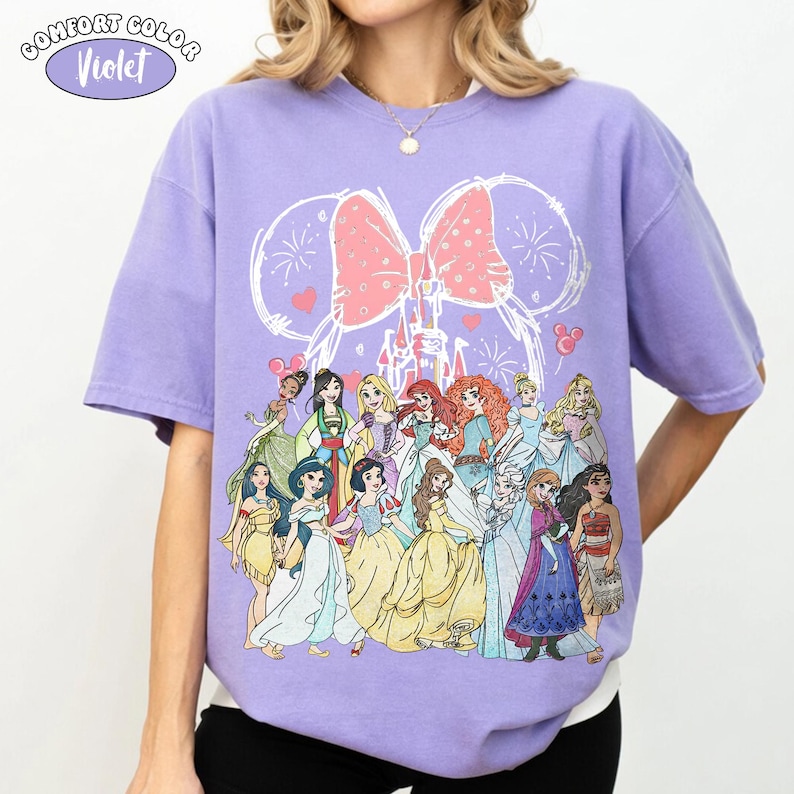 Op de afbeelding: Een violet Comfort Colors T-shirt met een afbeelding van Disney-prinsessen. Het ontwerp bevat een roze strik, vuurwerk en een kasteelomtrek. Het shirt heeft korte mouwen en een ronde hals.