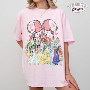 Op de afbeelding: Een lichtroze t-shirt met een afbeelding van Disney-prinsessen. Het ontwerp bevat een roze strik, kasteel en muisoren. De tekst "COMFORT COLOR Blossom" staat rechtsboven op de shirt.