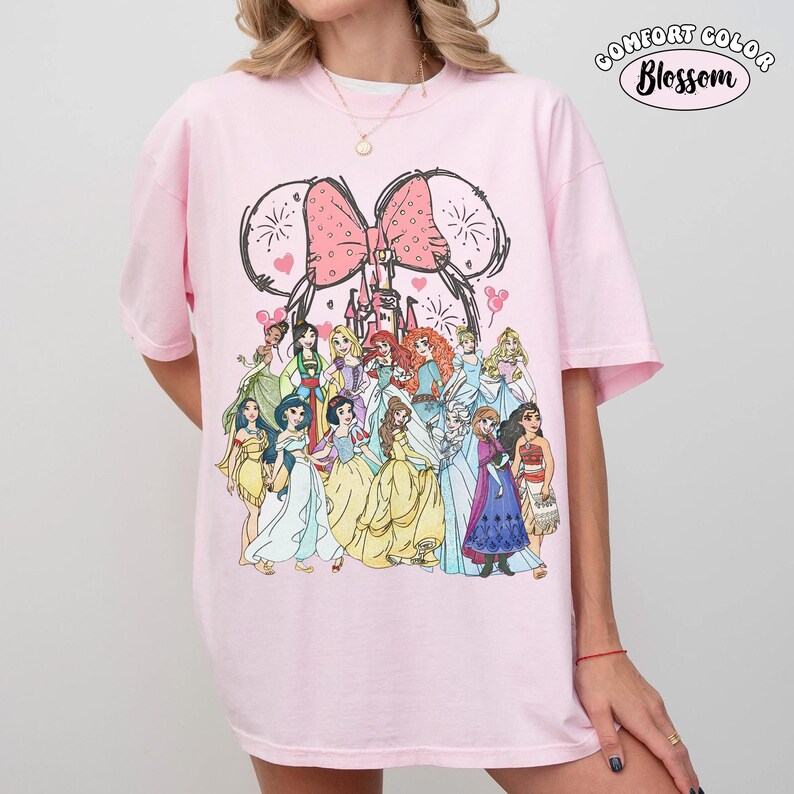 Op de afbeelding: Een lichtroze t-shirt met een afbeelding van Disney-prinsessen. Het ontwerp bevat een roze strik, kasteel en muisoren. De tekst "COMFORT COLOR Blossom" staat rechtsboven op de shirt.