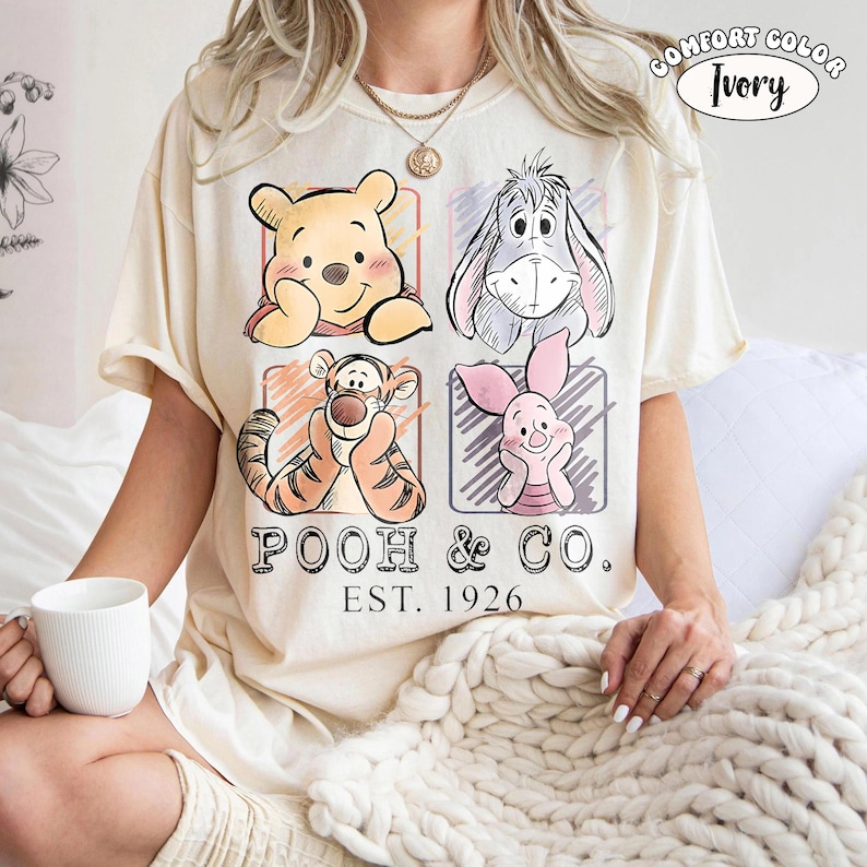 May include: Ivory-colored t-shirt featuring a graphic of Winnie the Pooh, Eeyore, Tigger, and Piglet. The shirt has the text "POOH & CO. EST. 1926" printed below the characters. The words "COMFORT COLOR Ivory" are printed at the top right.