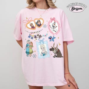 May include: Pink t-shirt featuring various characters and elements from the animated film "Frozen." The design includes framed portraits of Anna, Elsa, Olaf, and Kristoff, along with snowflakes and the word "FROZEN."