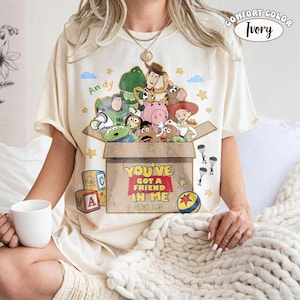 K&ouml;nnte beinhalten: Elfenbeinfarbenes Comfort Colors T-Shirt mit einem Toy Story-Motiv. Das Design zeigt Charaktere in einer Box mit dem Text "You've Got a Friend in Me Since 1995." Das Shirt hat auch den Namen "Andy" und Sterne.