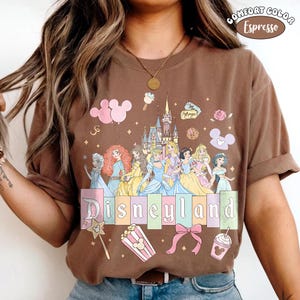 Puede incluir: Camiseta color espresso con un dise&ntilde;o de Disneyland. El gr&aacute;fico incluye princesas de Disney, un castillo y la palabra "Disneyland" en colores pastel. Elementos adicionales: palomitas de ma&iacute;z, una varita m&aacute;gica y un lazo.