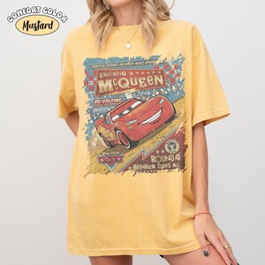 May include: Mustard-colored t-shirt featuring a graphic of Lightning McQueen from the Disney Pixar Cars movie. The design includes text that reads "Lightning McQueen" and "Winner Takes All". The shirt is a Comfort Color brand.