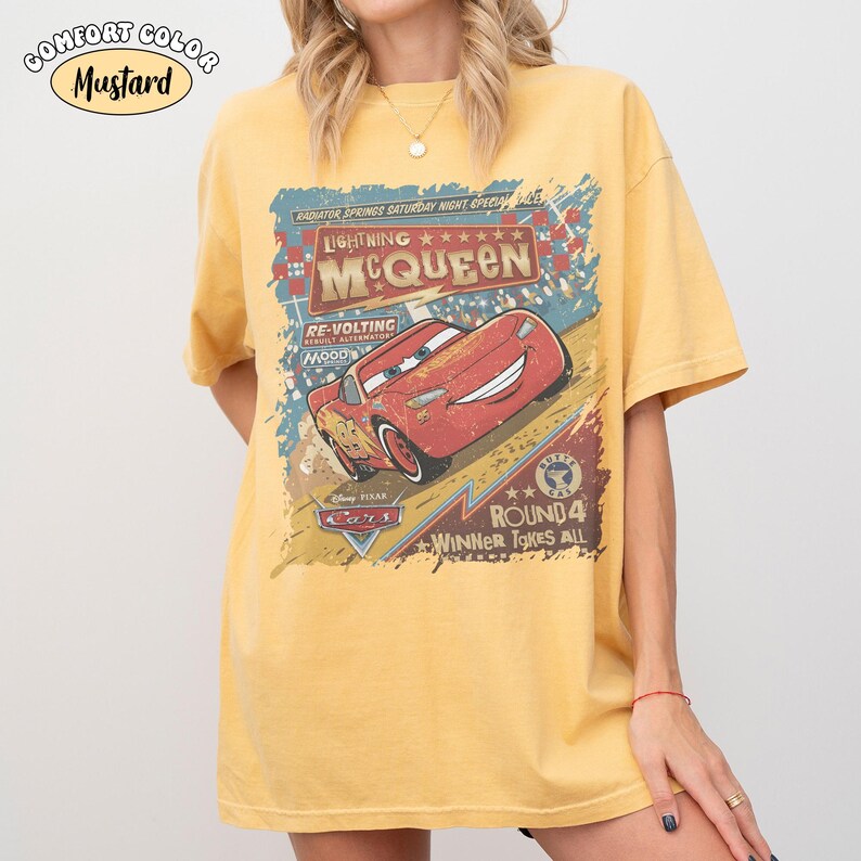 May include: Mustard-colored t-shirt featuring a graphic of Lightning McQueen from the Disney Pixar Cars movie. The design includes text that reads "Lightning McQueen" and "Winner Takes All". The shirt is a Comfort Color brand.