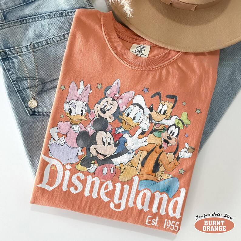 Pu&ograve; includere: Una t-shirt color arancione bruciato con una stampa grafica di Mickey Mouse, Minnie Mouse, Donald Duck, Daisy Duck, Goofy e Pluto. Il testo Disneyland Est. 1955 &egrave; stampato in bianco sotto i personaggi. La t-shirt ha un'etichetta Comfort Colors.