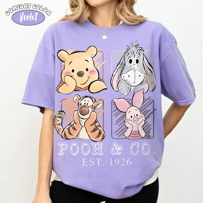 May include: A Comfort Colors violet t-shirt featuring Winnie the Pooh, Eeyore, Tigger, and Piglet. The graphic includes the text "POOH & CO. EST. 1926". The shirt has a relaxed fit and short sleeves.