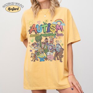 May include: A mustard yellow short-sleeve t-shirt featuring a graphic print with the text "AUTISM accept understand love" and various cartoon characters including a dinosaur, a space ranger, a cowboy, and a pig, along with a rainbow and sun.