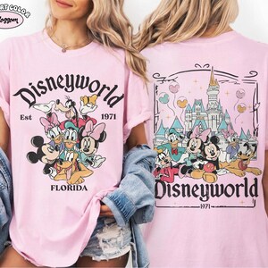 K&ouml;nnte beinhalten: Ein hellrosa T-Shirt mit einem Grafikdruck von Disney-Figuren wie Mickey, Minnie, Donald, Daisy, Goofy und Pluto. Die Vorderseite zeigt den Text Disneyworld Est 1971 Florida. Die R&uuml;ckseite zeigt das Schloss mit dem Text Disneyworld 1971.