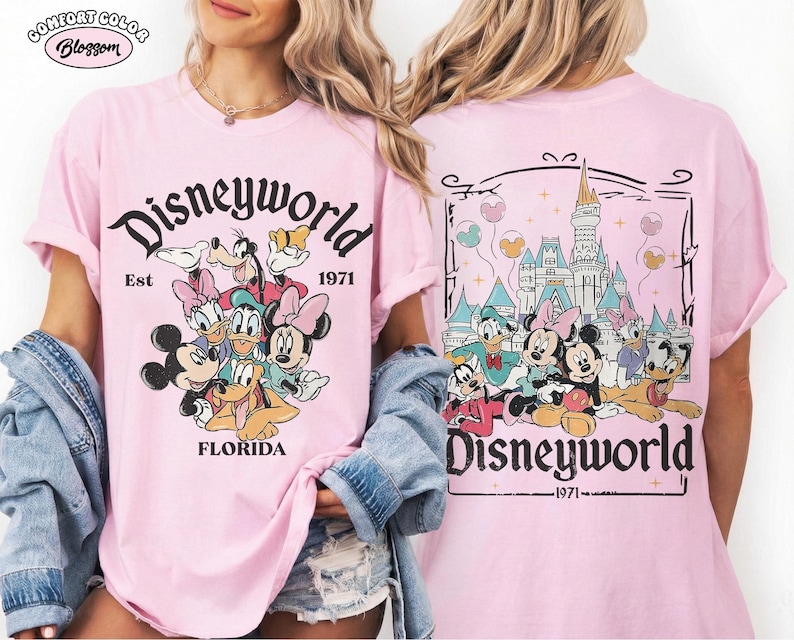 K&ouml;nnte beinhalten: Ein hellrosa T-Shirt mit einem Grafikdruck von Disney-Figuren wie Mickey, Minnie, Donald, Daisy, Goofy und Pluto. Die Vorderseite zeigt den Text Disneyworld Est 1971 Florida. Die R&uuml;ckseite zeigt das Schloss mit dem Text Disneyworld 1971.