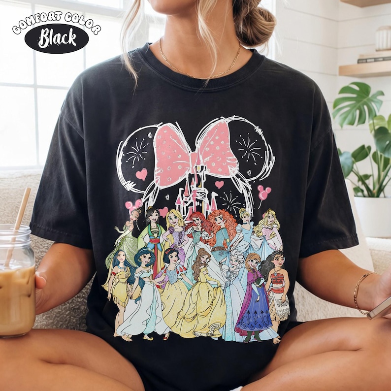 Op de afbeelding: Zwart Comfort Colors T-shirt met een Disney prinsessenafbeelding. Het ontwerp bevat een roze strik, kasteel en vuurwerk, met de tekst "Comfort Color Black" in een cirkel linksboven.