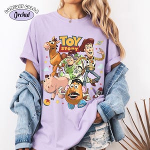 May include: A lilac Comfort Colors t-shirt featuring a graphic of characters from the animated film "Toy Story." The design includes Woody, Buzz Lightyear, Mr. Potato Head, Hamm, and Bullseye, with the film's logo. The shirt has short sleeves.