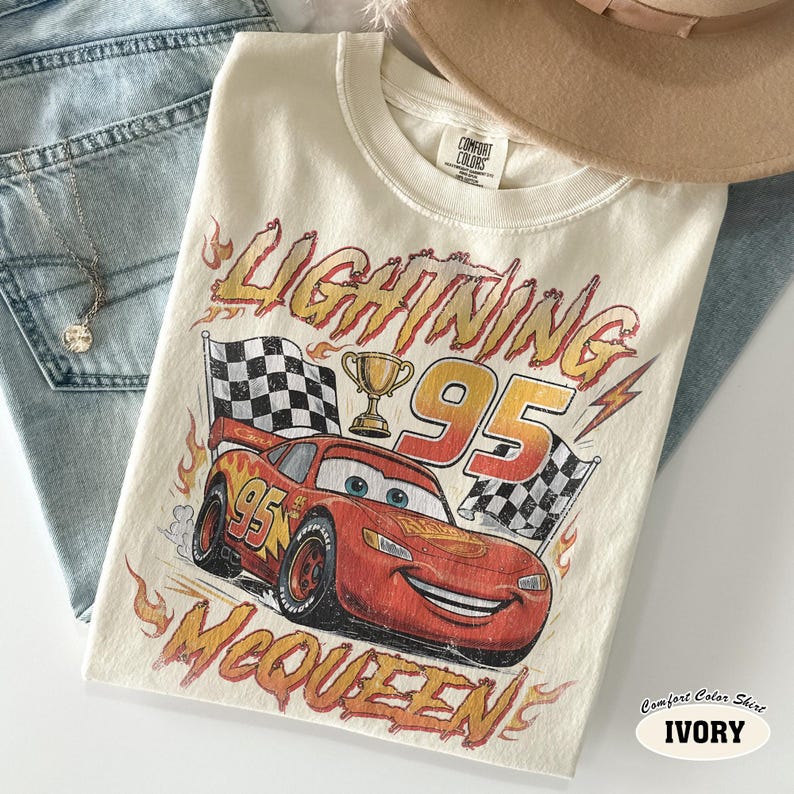 Puede incluir: Una camiseta color marfil con un gr&aacute;fico de un coche de carreras rojo con el n&uacute;mero 95, un trofeo dorado y banderas a cuadros. El texto dice LIGHTNING y MCQUEEN en una fuente naranja desgastada. Tiene cuello redondo y mangas cortas.