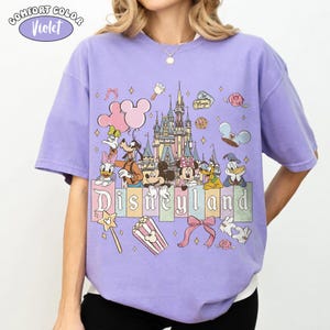 Peut inclure: T-shirt violet Comfort Colors avec un motif Disneyland. Le design comprend le ch&acirc;teau de Disneyland, Mickey Mouse, Minnie Mouse, Donald Duck, Dingo et d'autres personnages. Le mot "Disneyland" est affich&eacute; dans une police color&eacute;e.