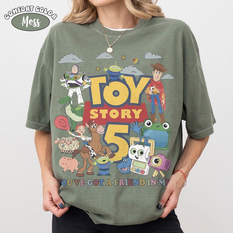 May include: A moss green short-sleeve t-shirt featuring a graphic print of Toy Story characters including Buzz Lightyear, Woody, Jessie, Rex, and others. The text reads TOY STORY 5 and YOU'VE GOT A FRIEND IN ME. A Comfort Color Moss label is at the top left.