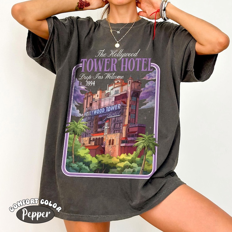 Op de afbeelding: Donkergrijs t-shirt met een afbeelding van het Hollywood Tower Hotel, palmbomen en de tekst "The Hollywood Tower Hotel Drop Ins Welcome 1994". Het shirt heeft een Comfort Color Pepper label.