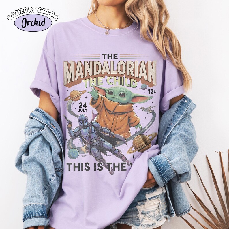 May include: An orchid-colored t-shirt featuring a graphic print of The Mandalorian and The Child in space. The text reads THE MANDALORIAN, THE CHILD, 24 JULY, 12c, and THIS IS THE WAY. The design includes planets, stars, and a spaceship.