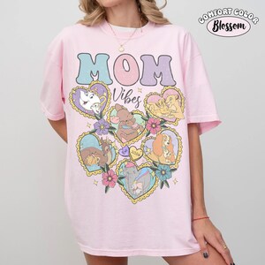 Pu&ograve; includere: Una maglietta Comfort Colors rosa chiaro con le parole "MOM Vibes" in un font retr&ograve;. La maglietta presenta illustrazioni a forma di cuore di personaggi Disney, tra cui Belle, Dumbo e Simba. La maglietta ha un girocollo e maniche corte.