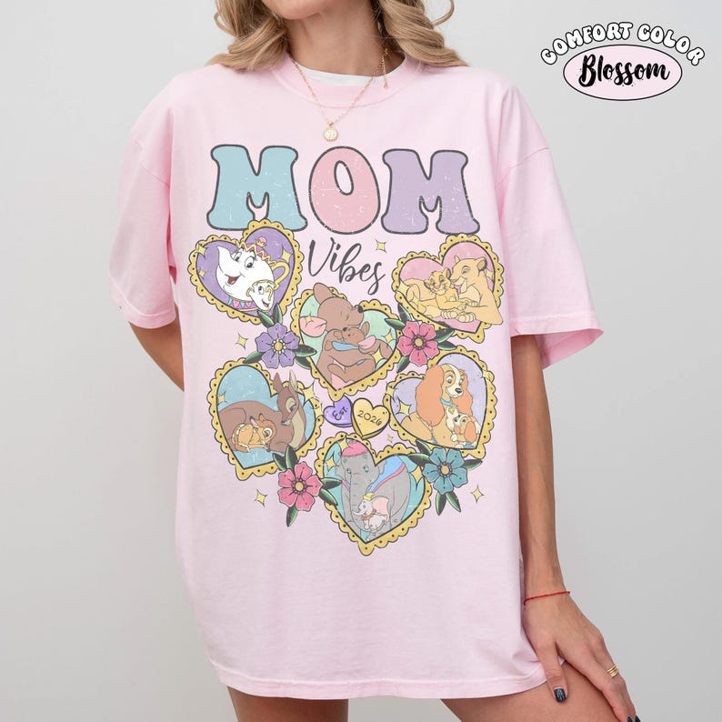 Pu&ograve; includere: Una maglietta Comfort Colors rosa chiaro con le parole "MOM Vibes" in un font retr&ograve;. La maglietta presenta illustrazioni a forma di cuore di personaggi Disney, tra cui Belle, Dumbo e Simba. La maglietta ha un girocollo e maniche corte.