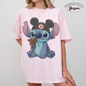 May include: A light pink t-shirt featuring a graphic of the character Stitch wearing a black ear hat with a red Mickey icon. Stitch is holding a brown Mickey-shaped ice cream bar. The text Comfort Color Blossom appears in the upper right corner.