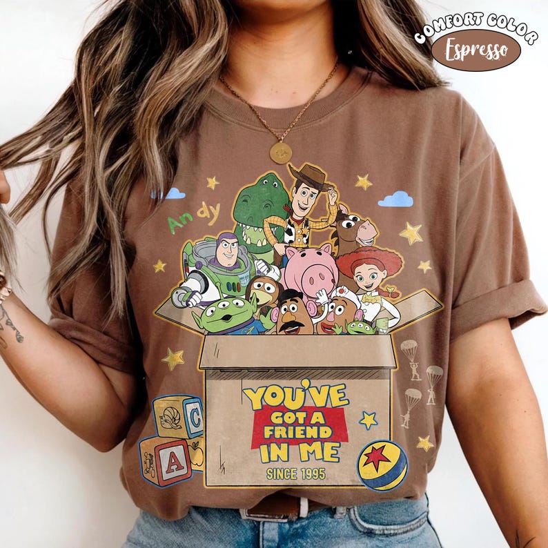 K&ouml;nnte beinhalten: Espressofarbenes T-Shirt mit einer Grafik von Toy Story-Charakteren in einer Box. Der Text "YOU'VE GOT A FRIEND IN ME SINCE 1995" steht darunter. Das Design enth&auml;lt den Namen "Andy" sowie Sterne und Wolken. Comfort Color.
