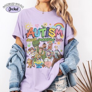 May include: A light purple t-shirt featuring a colourful graphic with the text "AUTISM accept understand love" and various cartoon characters including a cowboy, space ranger, dinosaur, and pig. The design includes a sun, rainbow, and puzzle pieces.